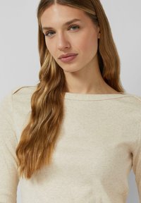 Beżowa prążkowana bluzka z długim rękawem, szerokim dekoltem w łódkę i subtelną fakturą. Włosy opadają luźnymi falami na jedno ramię. Odcień skóry jest neutralny.