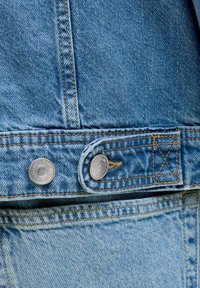 Záber zblízka na modrú rifľovú látku s dvoma striebornými kovovými gombíkmi a viditeľným oranžovým prešitím na bunde a rifle.