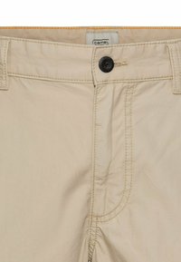 Beige broek met een gladde structuur, met een zwarte knoopsluiting, stiksels en een label met "camel" dat zichtbaar is op de tailleband.