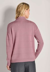 Helles rosa Strickpullover mit hohem Kragen, geripptem Saum und Streifenmuster auf der Rückseite. Kombiniert mit grauen Nadelstreifenhosen.