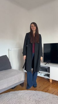 Μαύρο διπλόπατο παλτό με φαρδιά γιακά, φορεμένο πάνω από μπορντό πουλόβερ και μπλε καμπάνα τζιν, σε ένα μίνιμαλ σαλόνι.