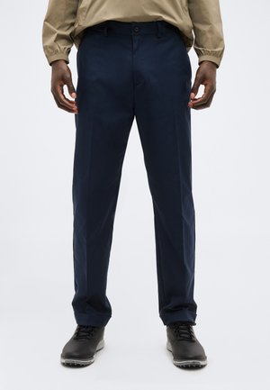 Homme portant un pantalon habillé bleu marine, des chaussures noires et une chemise beige à manches longues, debout devant un fond blanc.