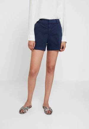 Vrouw draagt marineblauwe shorts, wit blouse met lange mouwen en open-toe sandalen met slangenhuidmotief, staand op een witte vloer.