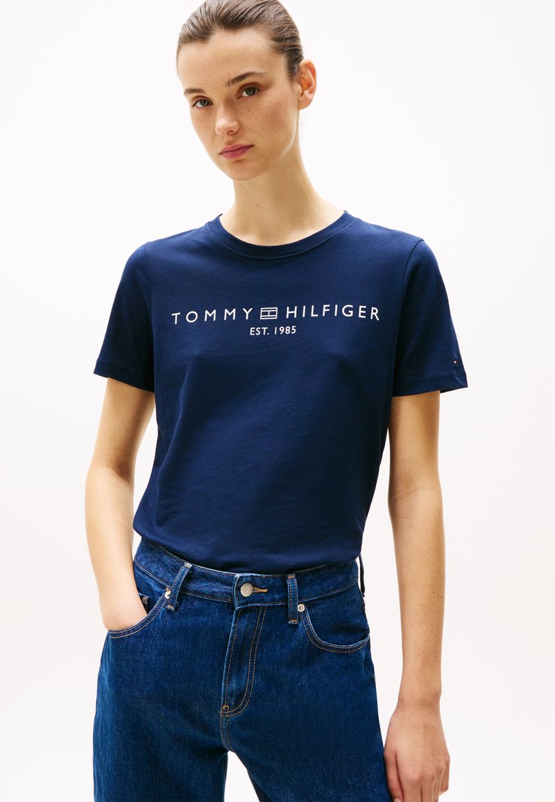 Tamsiai mėlyna medvilninė marškinėlių bluzonė su trumpomis rankovėmis ir balta "TOMMY HILFIGER EST. 1985" logo priekyje. Dėvima su aukšto juosmens džinsais.