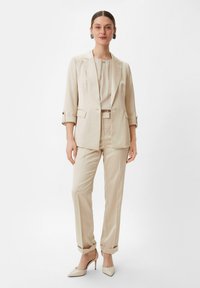 Beige tailored blazer and pants set made of smooth fabric. Features a single-button closure, rolled sleeves, and pointed-toe heels.