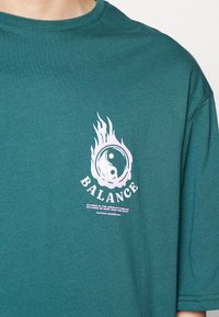 Tīrkīza kokvilnas T-krekls ar grafisko dizainu, kas attēlo liesmas un yin-yang simbolu. Zem tā balti ir rakstīts "BALANCE".