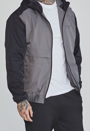 Homme portant une veste zippée grise et noire par-dessus un t-shirt blanc, avec des tatouages visibles au cou et aux mains, et un pantalon noir.