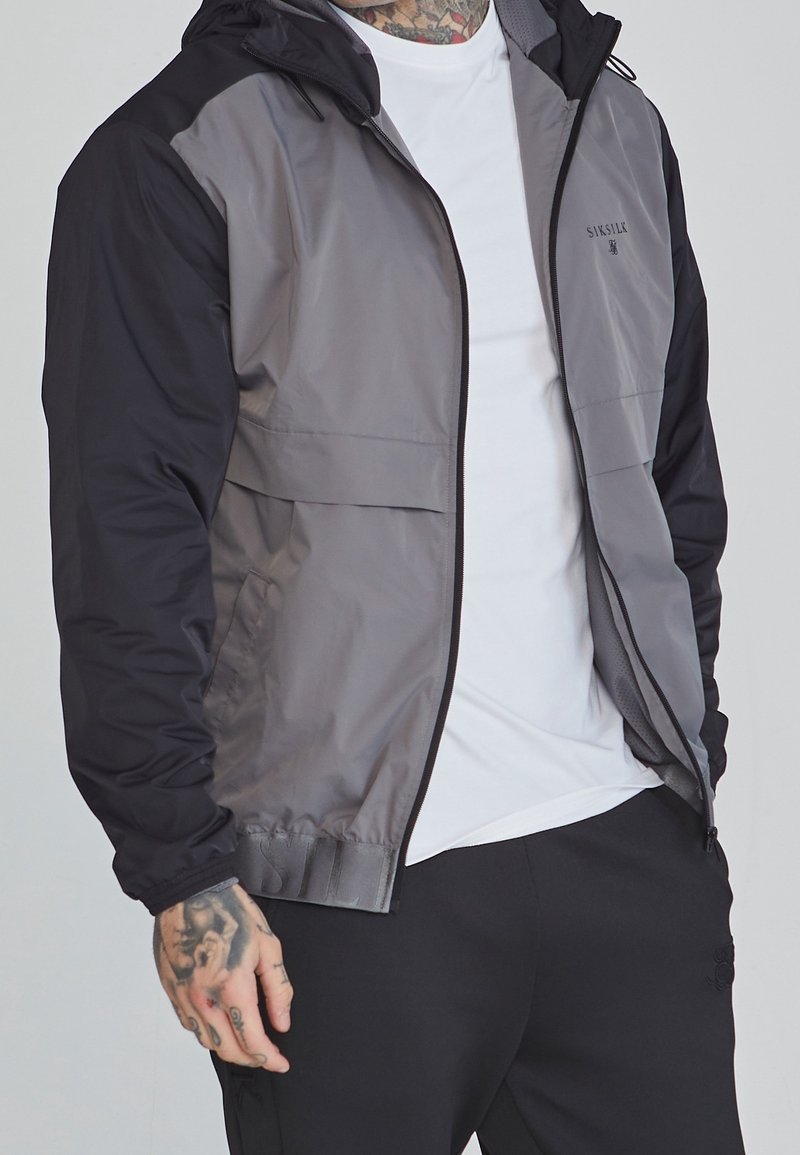 Homme portant une veste zippée grise et noire par-dessus un t-shirt blanc, avec des tatouages visibles au cou et aux mains, et un pantalon noir.