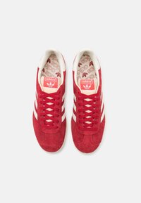 Baskets en daim rouge avec des bandes en cuir blanc, bout rond et lacets rouges. Logo iconique sur la languette et semelle intérieure à motif visible.