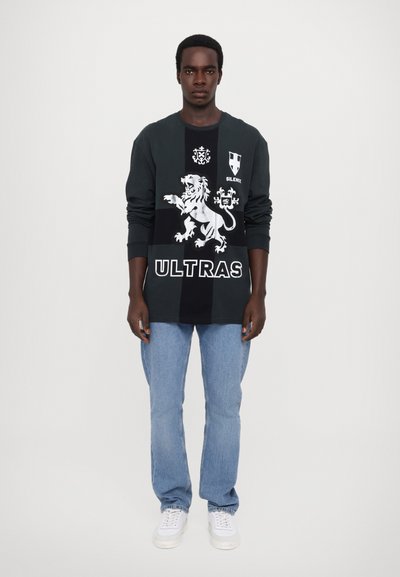 Sweat-shirt noir avec un graphique de lion, texte "ULTRAS" et un design de blason ; associé à un jean bleu clair et des baskets blanches. Fond uni.