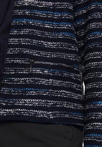 Gros plan sur un pull bleu marine texturé avec des rayures horizontales grises et bleues et une petite poche zippée noire.