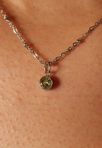 Zilveren ketting met een ronde, groene edelsteenhanger. De hanger is ingebed in een metalen omhulsel en rust tegen de huid met een gestructureerd oppervlakte.