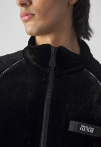 Close-up af en person iført en sort Versace Jeans Couture lynlåstrøje med høj krave, der viser en del af ansigtet og halsen.