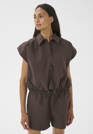 Femme aux longs cheveux foncés portant un haut sans manches marron à carreaux avec boutons et un short assorti, debout et regardant vers la gauche sur un fond blanc.
