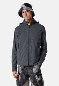 Grau Windbreaker-Jacke mit Kapuze, gelbem Reißverschlussdetail und zwei Seitentaschen. Kombiniert mit gemusterten schwarzen und grauen Cargo-Shorts.