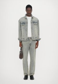 Light blue denim jacket with two chest pockets, worn over a white shirt, paired with matching jeans and black shoes, holding a brown bag.