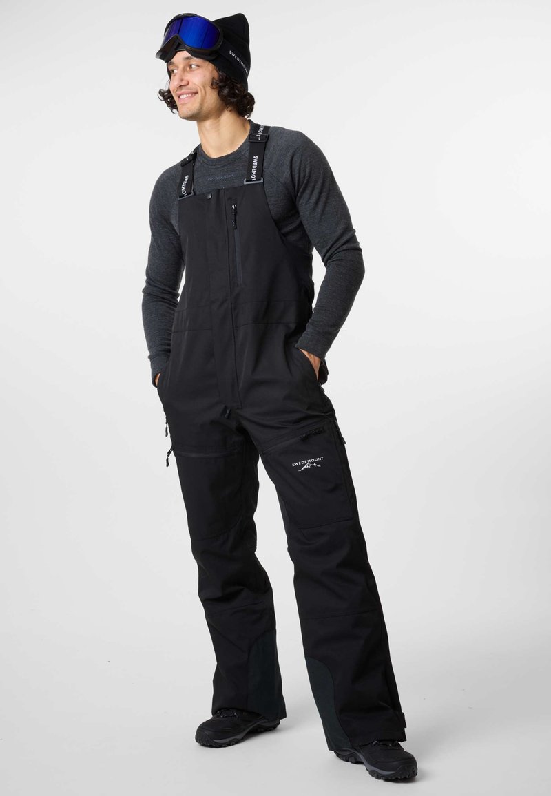 Black snow overalls with adjustable straps, zippered pockets, and reinforced knee patches. Worn over a grey long-sleeve shirt and black shoes.