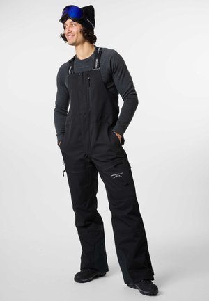 Schwarze Schneehosen mit verstellbaren Trägern, Reißverschluss-Taschen und verstärkten Kniebesätzen. Getragen über einem grauen Langarmshirt und schwarzen Schuhen.