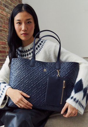 Femme aux longs cheveux foncés portant un pull blanc à motifs tient un grand sac fourre-tout bleu marine avec une pochette zippée assortie.