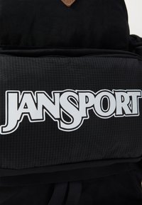 Zwarte Jansport rugzak met een gestructureerde stof en een gestreept patroon, opvallend wit logo en een bruin leren accent aan de bovenkant.