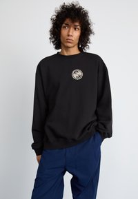 Sudadera negra hecha de una tela suave con un diseño gráfico circular color crema en el pecho. Estilo holgado con puños y dobladillo acanalados.
