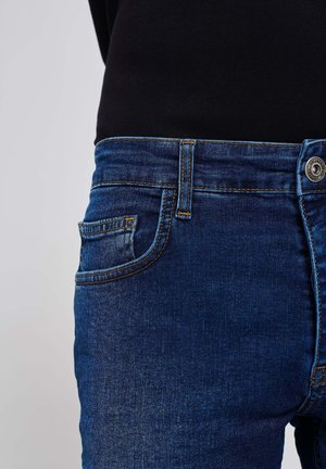 Jeans in denim blu con una vestibilità standard, dotati di una tasca frontale, passanti per cintura e chiusura con bottone in metallo. La texture appare liscia e tessuta.