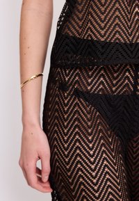 Abito di pizzo nero con un motivo a zigzag, texture trasparente e design aderente. Include un sottile braccialetto d'oro al polso.