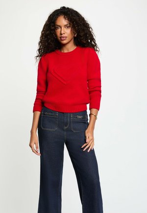 Mujer con cabello rizado, vistiendo un suéter rojo con diseño de corazón y jeans de pierna ancha azul oscuro, de pie contra un fondo blanco.