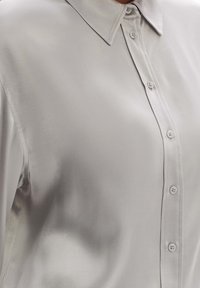 Chemisier gris clair en tissu lisse et réfléchissant. Il dispose d'un col classique, d'une patte de boutonnage sur le devant et de boutons ressemblant à des perles.