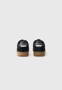 Black suede sneakers with a gum rubber outsole, featuring a white logo tag on the heel and a low-top design.