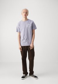 Camiseta de algodón de color púrpura claro con un gráfico redondo en el pecho, combinada con pantalones marrones oscuros y zapatillas con estampado.