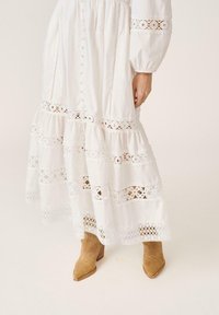 Robe longue blanche avec panneaux en dentelle et détails à boutons, associée à des bottines en daim marron clair à talon empilé sur un fond uni.