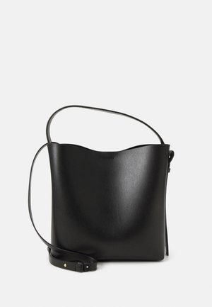 Musta nahkainen hoitolaukku, jossa säädettävä olkahihna, sileä pinta ja minimalistinen muotoilu vaalealla taustalla.