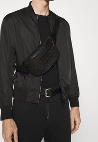 Černá bomber bunda přes přiléhavé černé tričko, doplněná o texturovanou černou ledvinku a černé kalhoty s jemným vzorem.