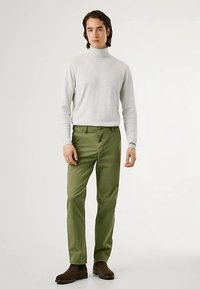 Camisola cinza de gola alta, calças verde-oliva, textura lisa, design justo, botas castanhas até ao tornozelo, com logótipo na manga esquerda.