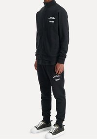 Černá zipová bunda a odpovídající tepláky s textem "V ZLATĚ DŮVERUJEME". Tenisky mají černobílý design se zlatými akcenty.