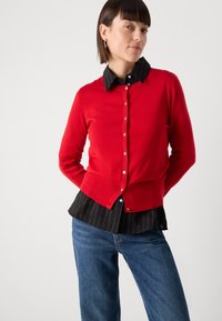 Red knit cardigan with a round neckline, button closure, and ribbed hem. Worn over a black and white striped shirt with a collar. Blue jeans.
