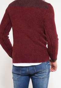 Bordeaux gestrickter Pullover mit strukturiertem Stoff, gerippten Bündchen und Saum, getragen über Jeans, zeigt die Rückansicht und Farbverlauf der Schulter.