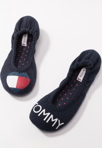 Chaussons de ballet Tommy Hilfiger bleu marine avec bords élastiques, semelles intérieures à motif d'étoiles rouges, logo en forme de cœur sur la chaussure gauche, et inscription "TOMMY" sur la droite.