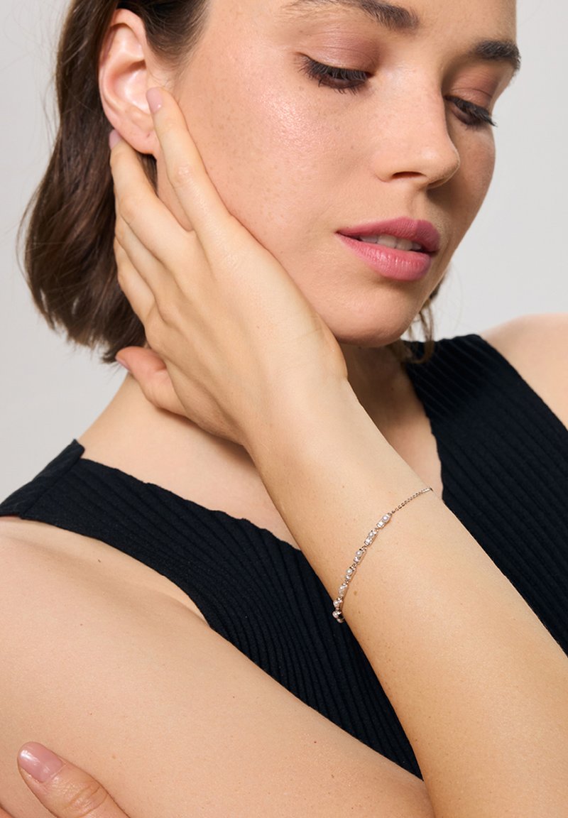 Srebrna bransoletka z małymi, okrągłymi, przezroczystymi kamieniami osadzonymi w delikatnym łańcuszku, noszona na nadgarstku w zestawieniu z teksturowanym czarnym topem.