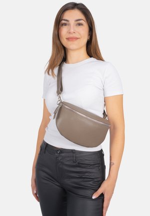 Crossbody-Tasche aus strukturiertem taupefarbenem Leder in runder Form, mit Reißverschluss und verstellbarem Tragegurt. Getragen über einem weißen T-Shirt.