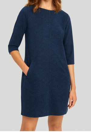 Vrouw draagt een knielange, marineblauwe jurk met driekwarts mouwen en zijzakken, staand tegen een lichtgrijze achtergrond.