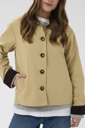 Femeie purtând o haină bej cu nasturi și manșete striate maro închis, peste un pulover tricotat gri și alb, asortată cu blugi albaștri.