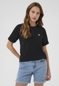 T-shirt en coton noir avec des manches retroussées et un petit emblème en forme de cœur blanc sur la poitrine gauche, associé à un short en denim bleu plié.