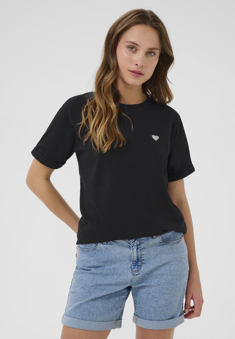 T-shirt en coton noir avec des manches retroussées et un petit emblème en forme de cœur blanc sur la poitrine gauche, associé à un short en denim bleu plié.