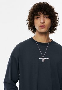 Maglietta a maniche lunghe blu scuro con logo bianco "STWD STUDIO" sul petto. Collana d'argento con pendente. Capelli ricci.