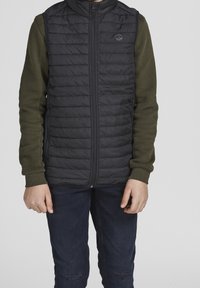 Person wearing black quilted vest over olive green long-sleeve shirt and dark blue jeans, standing against plain light background.