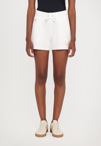 Pantalones cortos blancos con cintura elástica y cordón, con un logo en la pierna izquierda, combinados con zapatillas de color beige claro con suelas de goma.