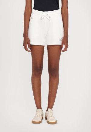 Shorts bianchi con vita elastica e cordoncino, con un logo sulla gamba sinistra, abbinati a sneakers beige chiaro con suola in gomma.