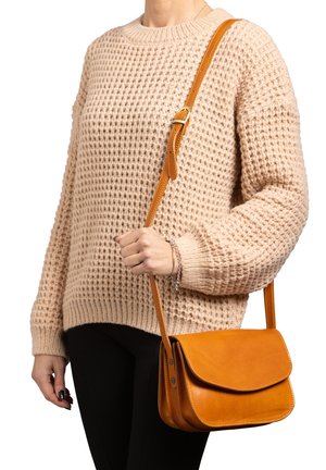 Personne portant un pull beige texturé et un pantalon noir, tenant un sac bandoulière en cuir jaune moutarde par sa sangle réglable.